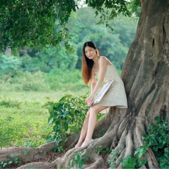 A woman in a light dress sits on the large roots of a tree in a grassy, green outdoor setting, holding a book and looking at the camera.
