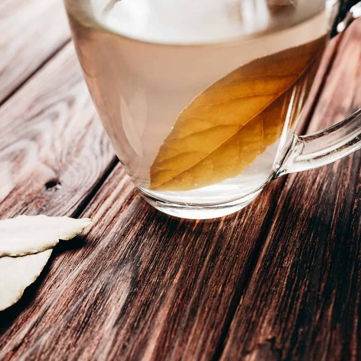 A clear glass mug of bay leaf tea with a bay leaf inside sits on a wooden surface, with two dried bay leaves lying nearby.