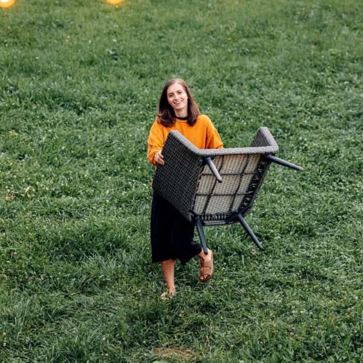 A woman wearing an orange sweater and black skirt carries an upside-down outdoor chair across a grassy yard.