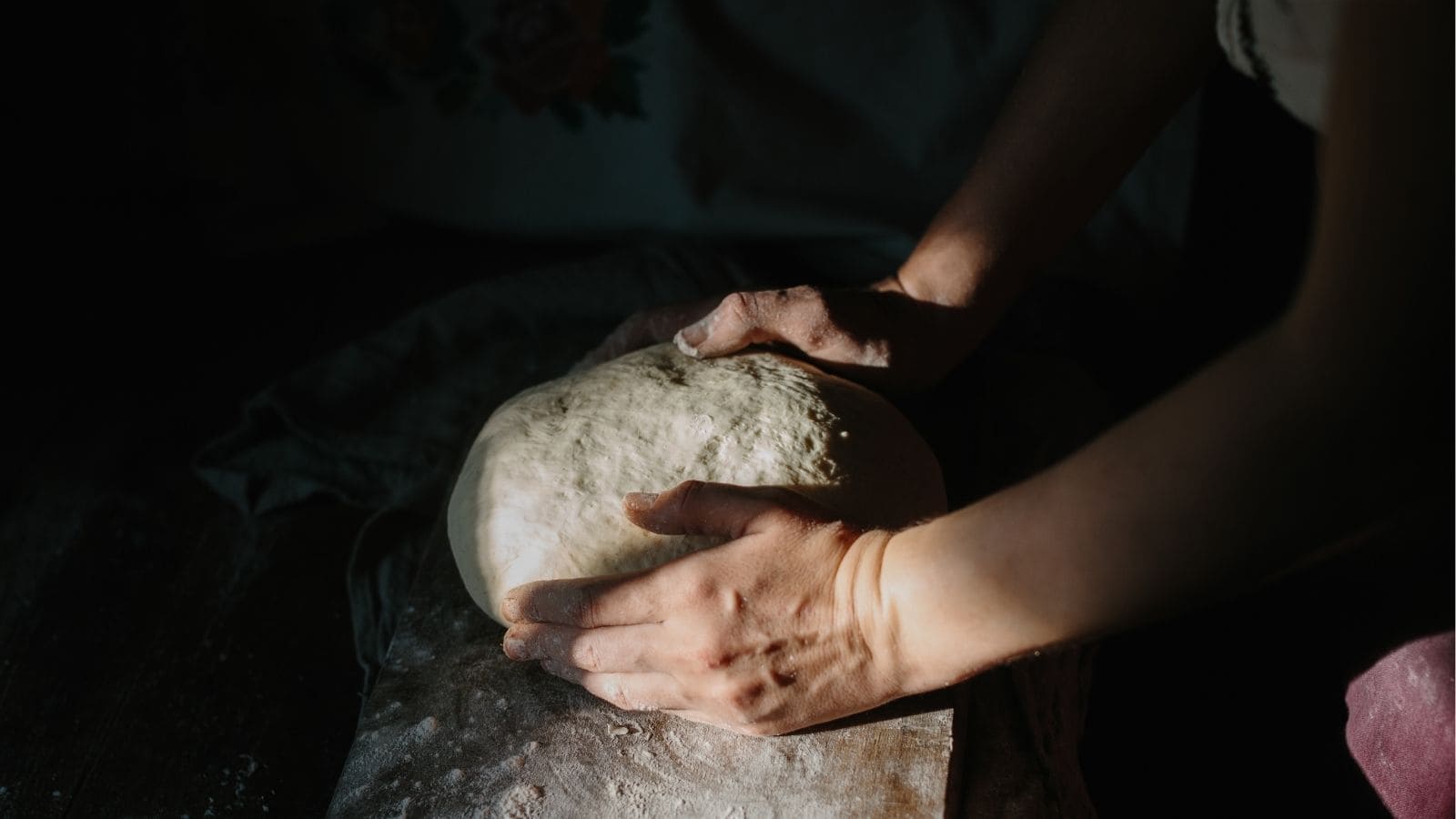 Hands kneading dough on a floured surface with soft lighting, partially illuminating the scene.