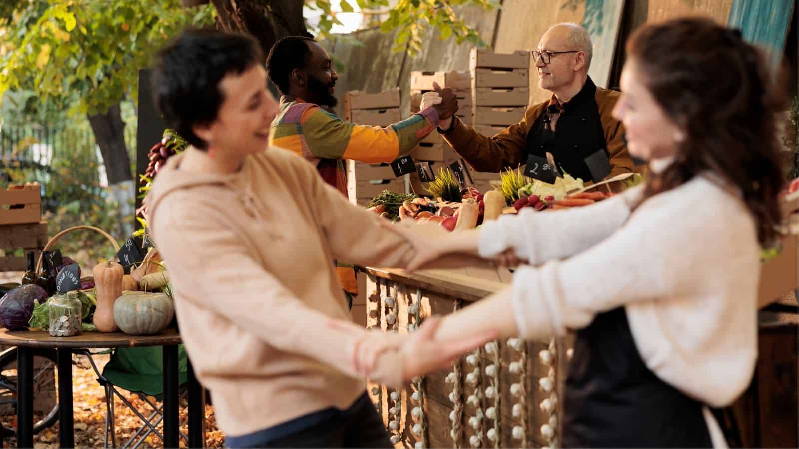 Two people hold hands and smile in the foreground, while two others greet each other with a handshake at a market stall with vegetables in the background.