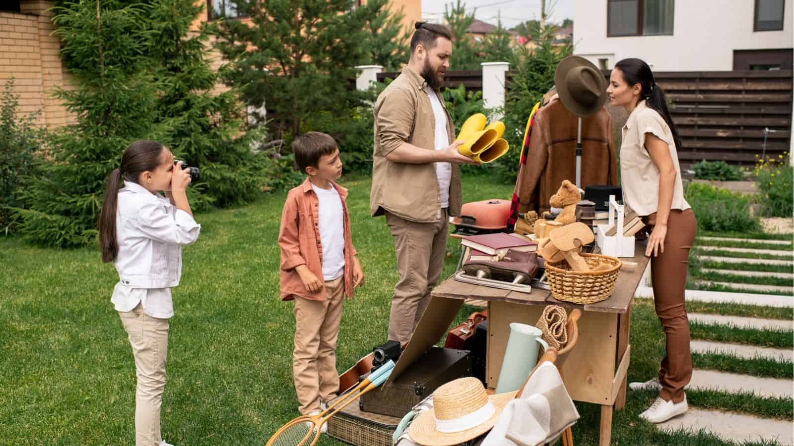 A family stands in a backyard by a table with various items for sale; the girl takes a photo while others examine and arrange objects.