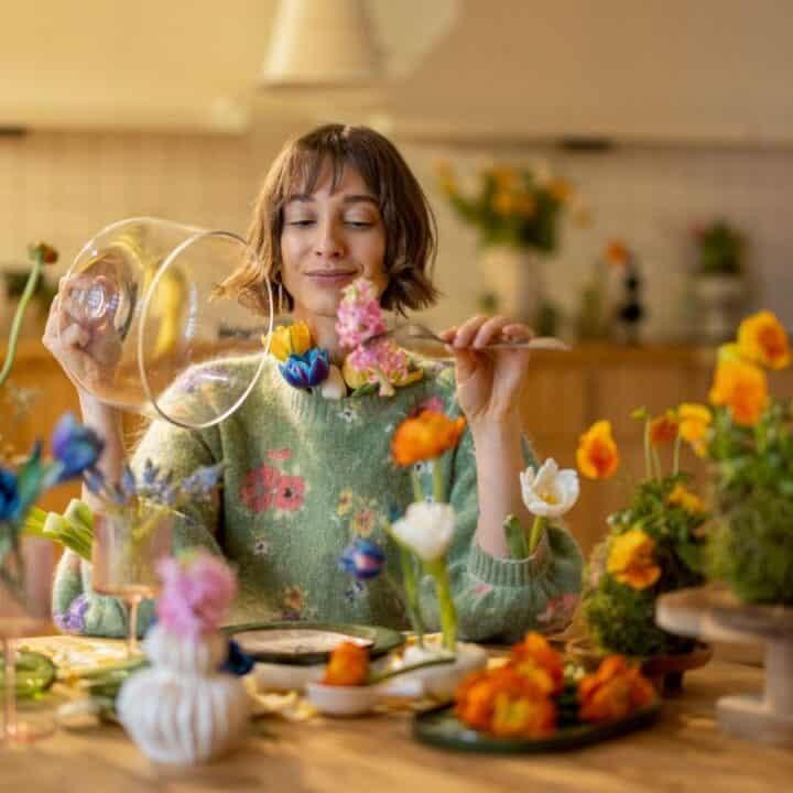 A woman arranges colorful flowers in vases on a wooden table in a kitchen, holding a glass container in one hand.