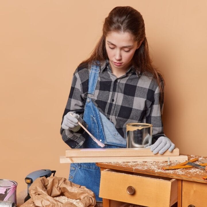 A young person in plaid shirt and overalls applies paint or varnish to a wooden plank at a workbench with various tools and supplies.