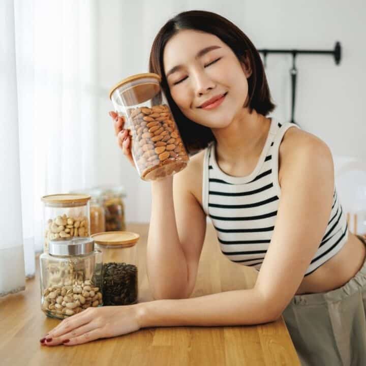 A woman in a striped tank top smiles while holding a jar of almonds in a kitchen. Several other jars with nuts and seeds are on the counter.
