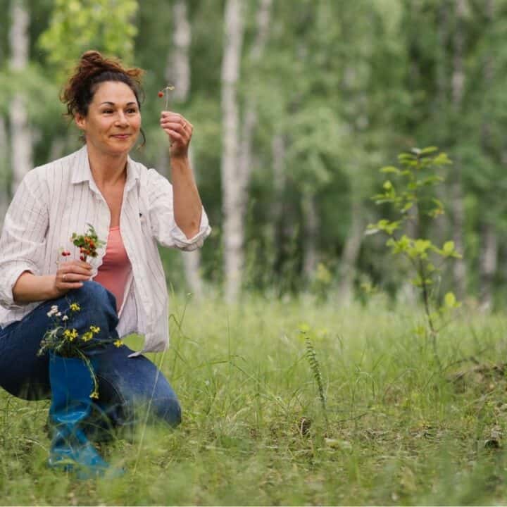 A woman kneels on grass in a forest, holding and examining wildflowers, with trees and greenery in the background.