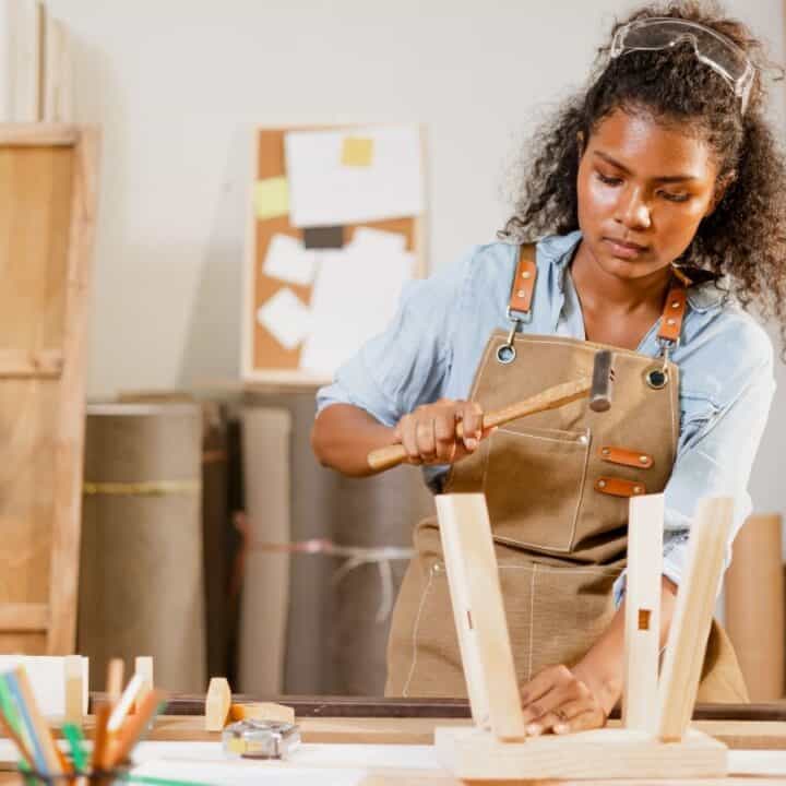 A woman wearing an apron and safety glasses uses a hammer to assemble a wooden piece at a workbench in a carpentry workshop.