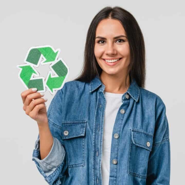 Woman in a denim shirt smiling and holding a paper cutout of a green recycling symbol against a plain light background.