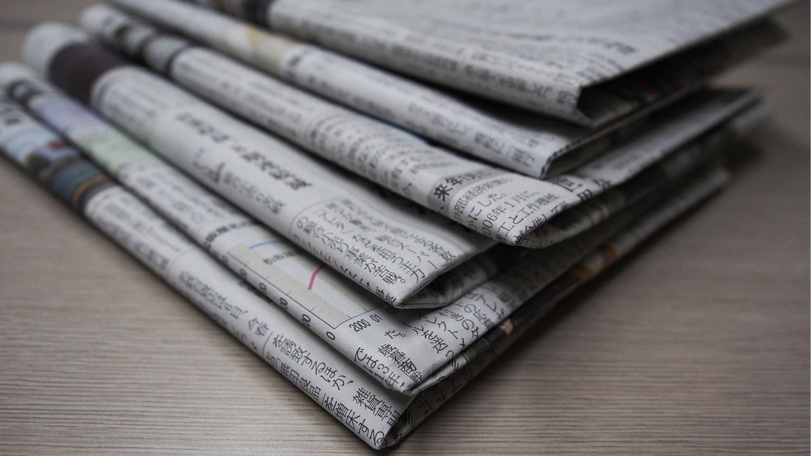 A stack of folded newspapers with Japanese text lies on a light-colored wooden surface.