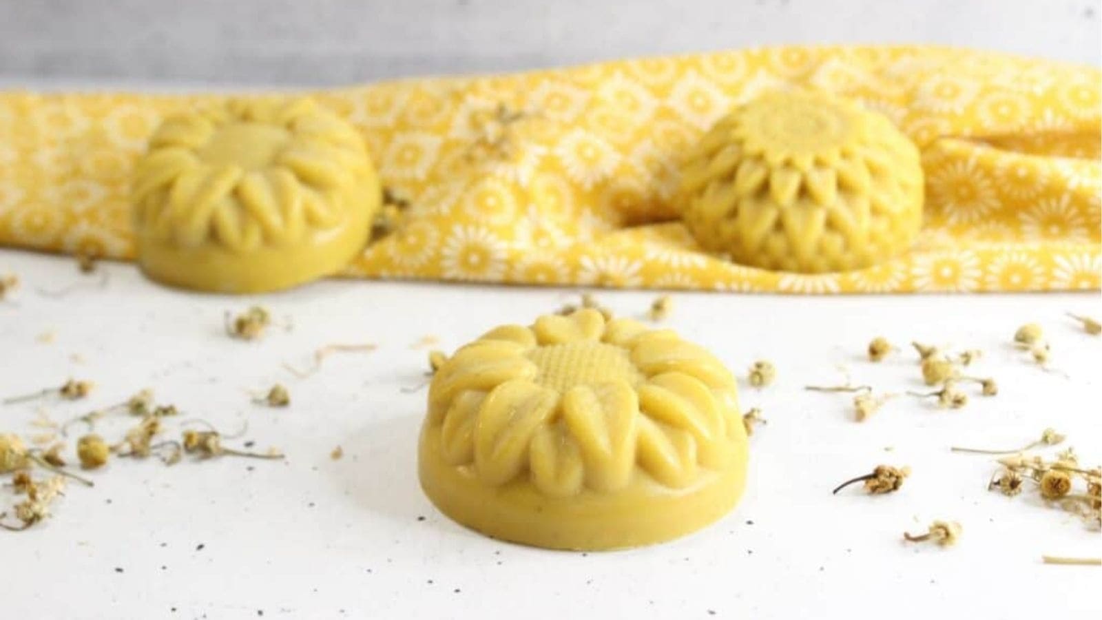 Yellow, flower-shaped solid lotion bars are placed on a white surface with dried flowers and a yellow patterned cloth in the background.