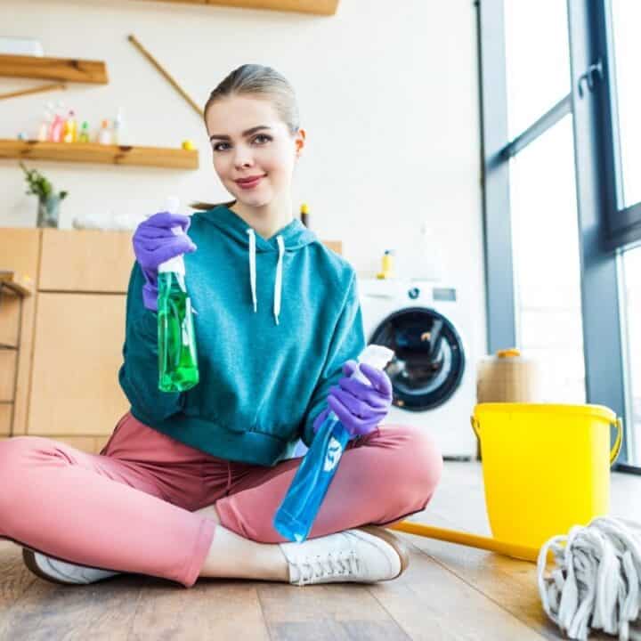 A woman wearing gloves sits cross-legged on the floor with cleaning supplies, including spray bottles, a bucket, and a mop, in a modern, well-lit room.