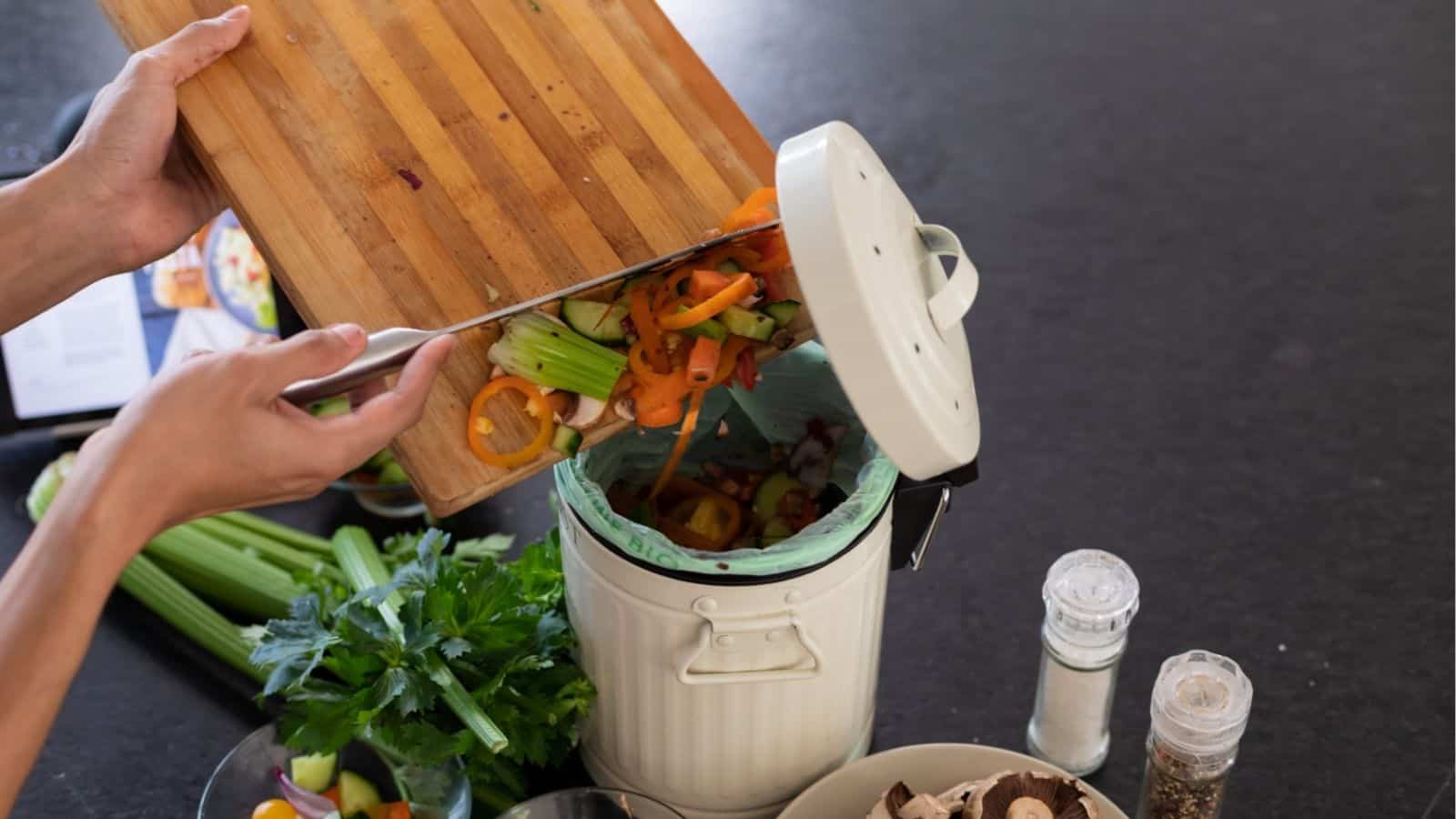 A person transfers vegetable scraps from a cutting board into a kitchen compost bin; celery, bell peppers, and mushrooms are visible on the counter.