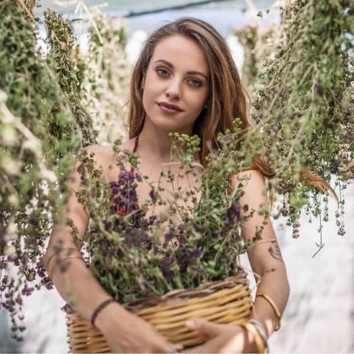 A woman holding a basket filled with herbs stands among hanging bunches of dried plants, looking directly at the camera.