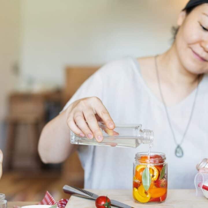 A person pours liquid into a jar of sliced vegetables at a kitchen counter with a basket of produce nearby.