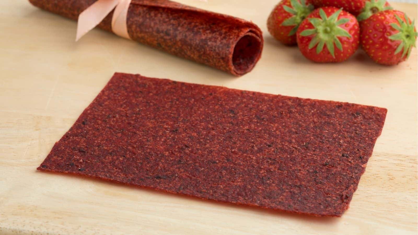 A sheet of strawberry fruit leather lies on a wooden surface, with fresh strawberries and a rolled-up piece of fruit leather in the background.