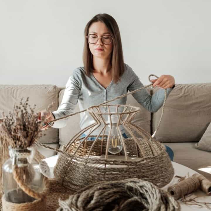 A woman sits on a sofa and wraps rope around a metal lampshade frame at a table with craft supplies in a living room.