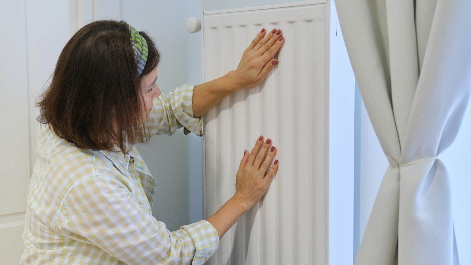 A woman wearing a plaid shirt touches a white radiator with both hands near a light-colored curtain.
