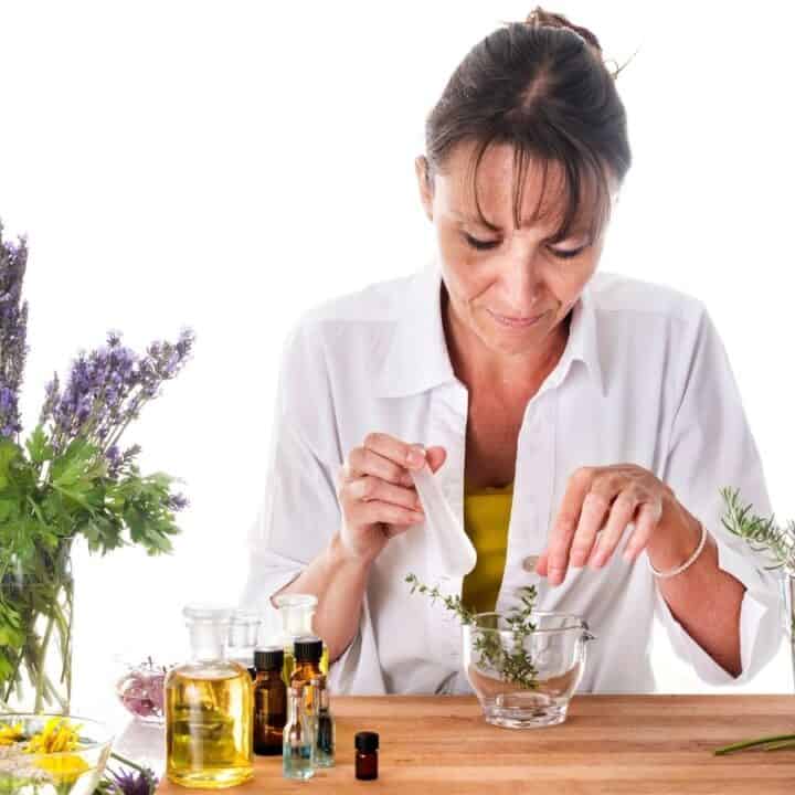 A woman in a white shirt prepares herbal extracts at a table with various plants, flowers, and bottles of oil on a white background.