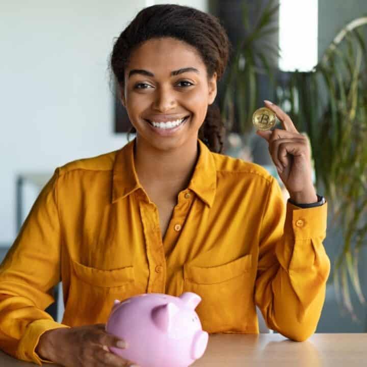Woman in a yellow blouse sits at a table, holding a coin in one hand and a pink piggy bank in the other, smiling at the camera.
