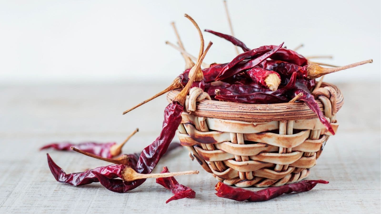 A small woven basket filled with dried red chili peppers sits on a light wooden surface, with some peppers scattered beside it.