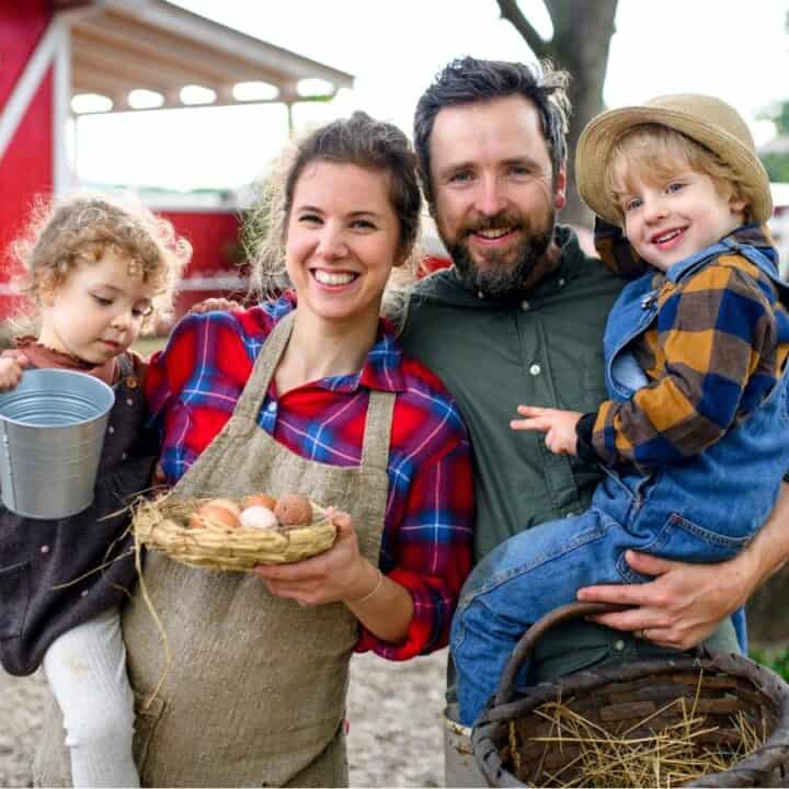 A family of four stands outside on a farm, smiling. The mother holds eggs in a nest, the father holds a young boy, and a young girl holds a metal bucket.