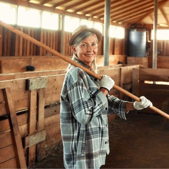 Older woman wearing gloves and a plaid shirt stands inside a barn holding a rake over her shoulder, with hay and wooden stalls visible in the background.