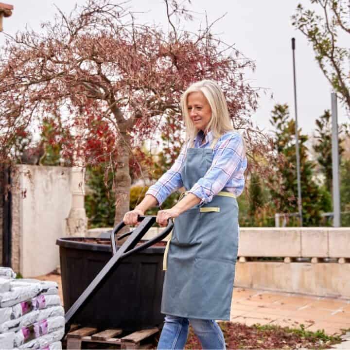 A woman wearing an apron uses a hand truck to move bags of soil or mulch in an outdoor garden center.