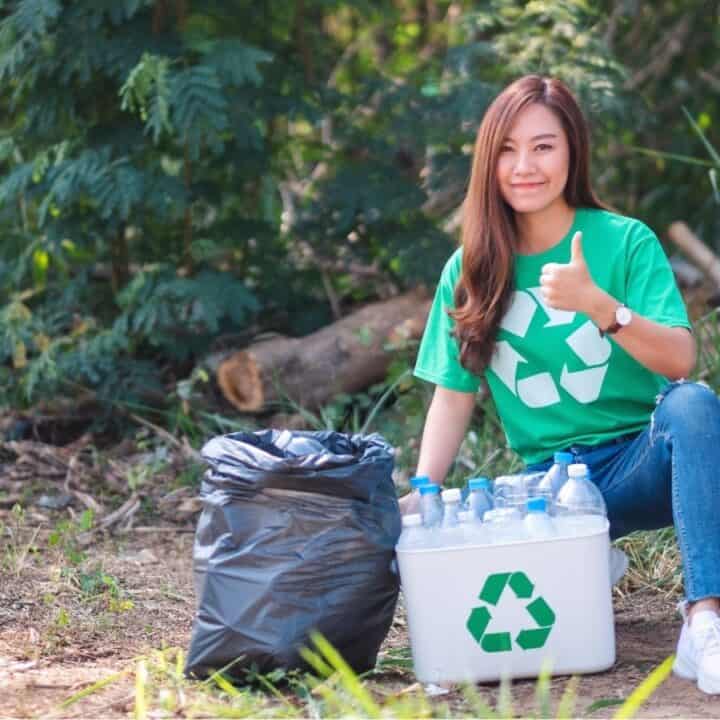 A woman wearing a green recycling shirt sits outdoors, giving a thumbs up beside a trash bag and a plastic bin filled with bottles.