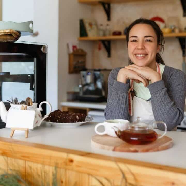 A woman wearing an apron stands behind a café counter with a teapot, cup, and cake on display, smiling at the camera.