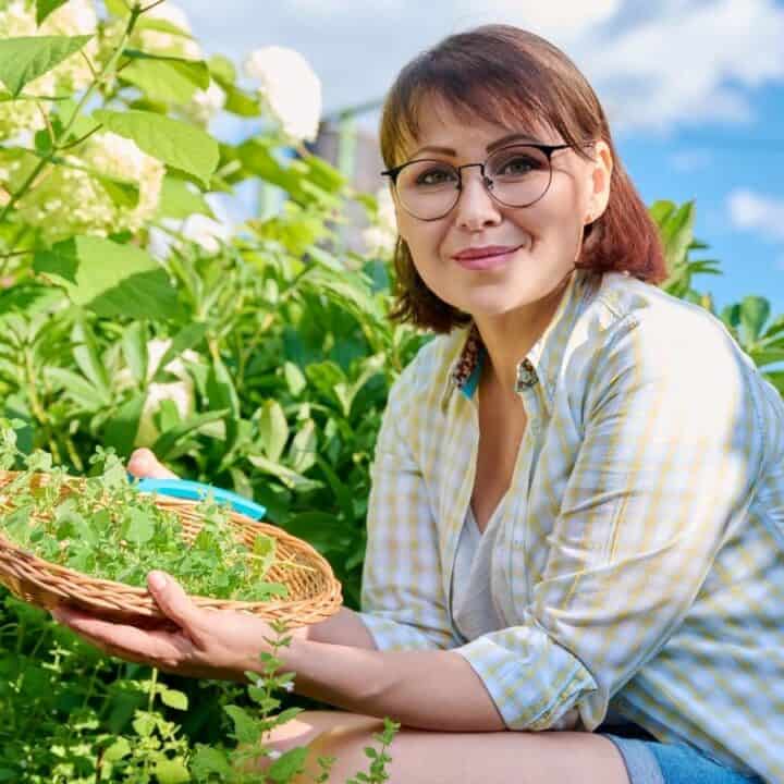 A woman wearing glasses sits in a garden, holding a basket of freshly picked herbs, surrounded by green plants and flowers under a blue sky.