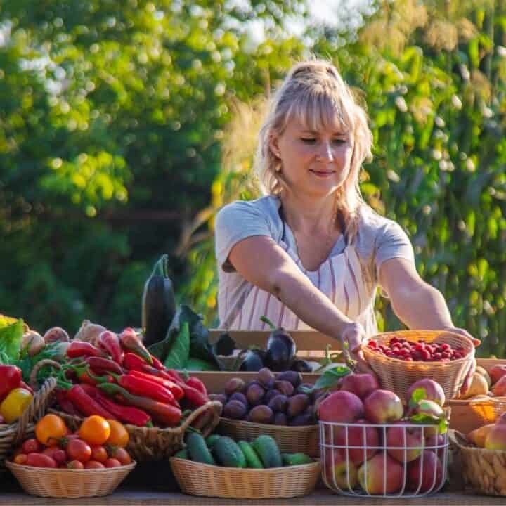 A woman arranges baskets of fresh fruits and vegetables on an outdoor table at a market stand, with greenery and crops in the background.