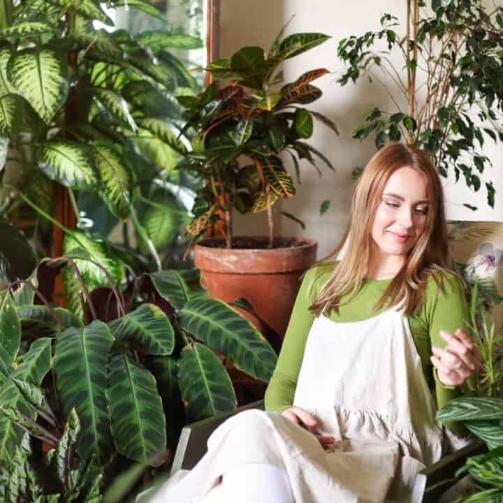 A woman sits on a chair surrounded by various potted houseplants in a well-lit room, wearing a green shirt and white overalls.