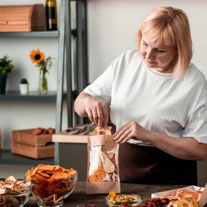 A woman packing dried fruit snacks into paper bags at a kitchen counter with jars and packets of snacks around her.