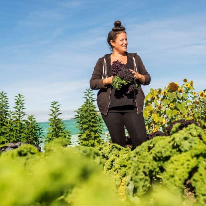 A woman stands in a garden holding freshly picked greens, surrounded by various plants and sunflowers under a blue sky.
