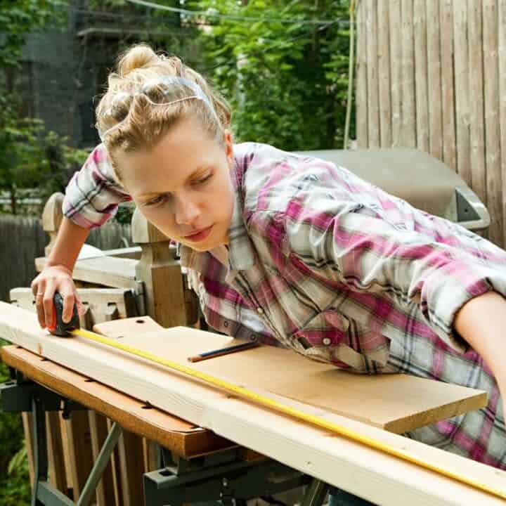 A person in a plaid shirt measures a wooden board with a tape measure on an outdoor workbench.