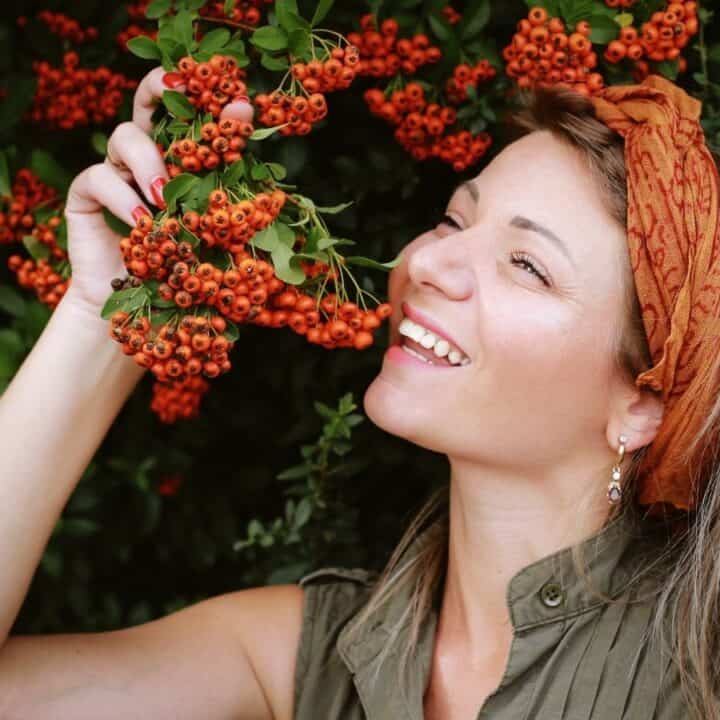 A woman with long hair and an orange headband smiles while holding a branch of orange berries near her face. Green foliage fills the background.