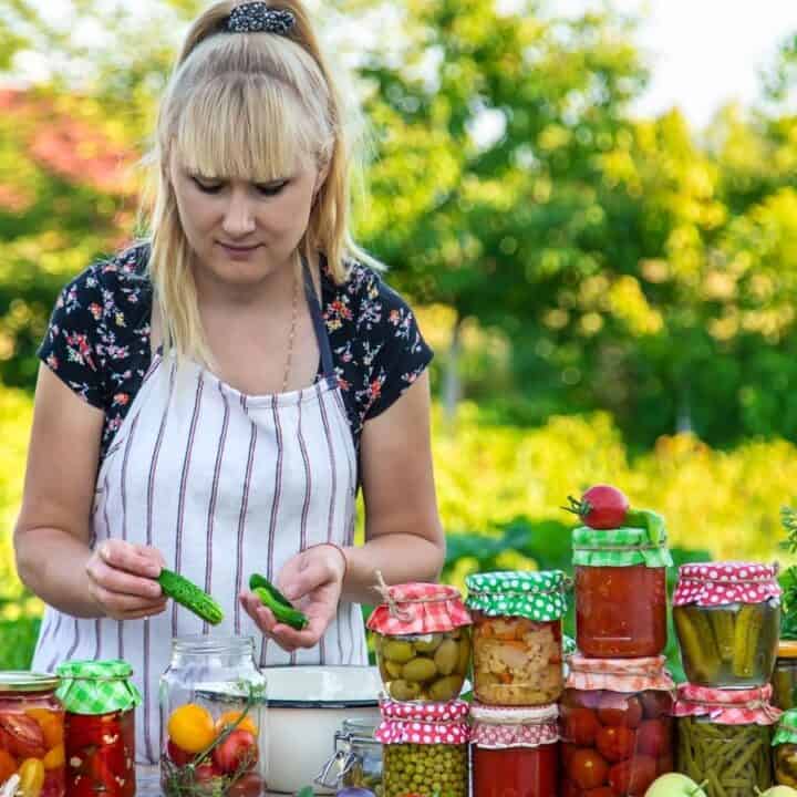 A woman in an apron is canning vegetables outdoors, surrounded by jars of preserved produce and fresh vegetables on a table.