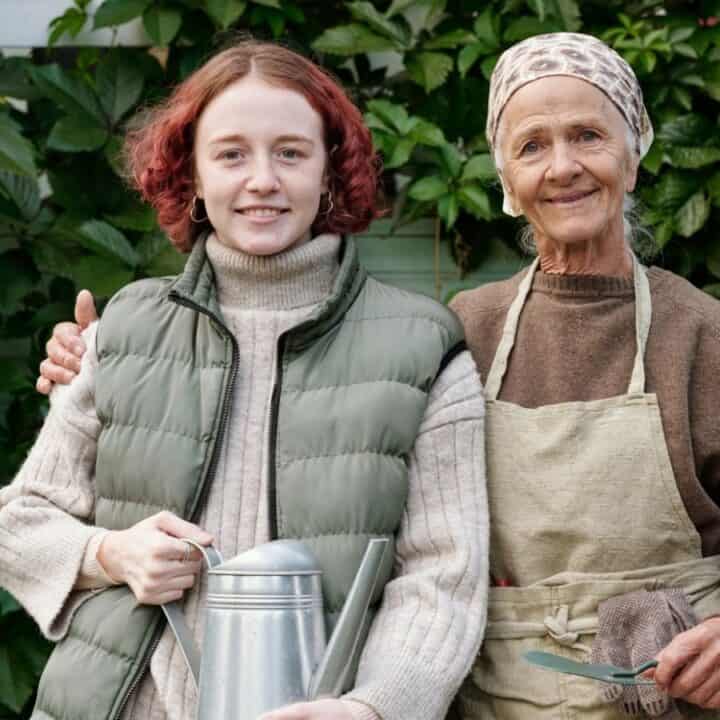 Two women stand in front of green foliage. One holds a watering can and wears a vest; the other wears an apron and headscarf, holding gardening tools. They are both smiling.