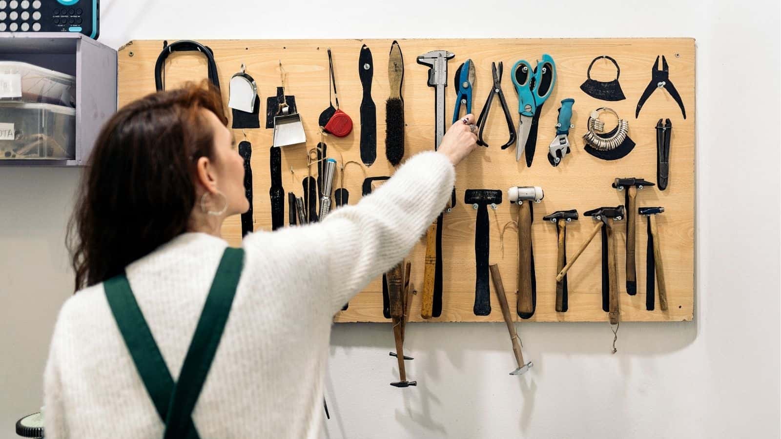 A person in a white sweater reaches for pliers on a wall-mounted tool board organized with various hand tools.
