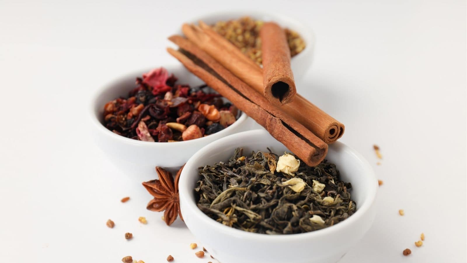 Three white bowls containing dried tea leaves, dried fruit pieces, and mixed spices, with cinnamon sticks and star anise on a white background.