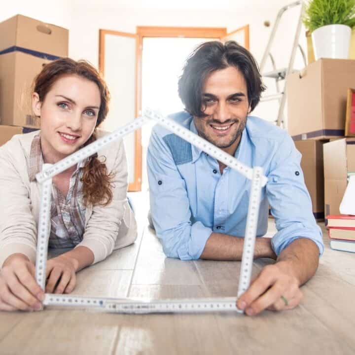 A woman and a man lying on the floor, smiling and holding a folding ruler shaped like a house, surrounded by moving boxes and household items.