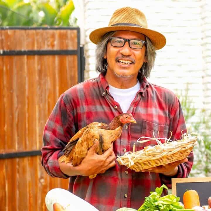 A man wearing a hat and glasses holds a brown chicken and a wicker basket, standing near a table with vegetables in an outdoor setting.