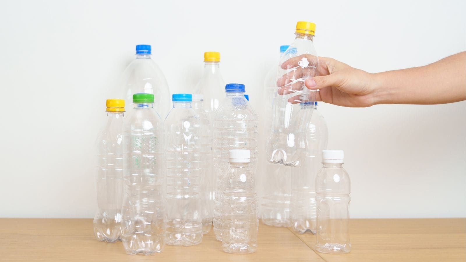 A hand holds one of several empty clear plastic bottles with various colored caps arranged on a wooden surface against a plain white background.