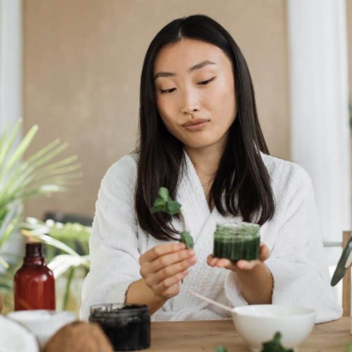 A woman in a white robe sits at a table with natural skincare products, holding a green jar and a sprig of mint, surrounded by plants and fruit.