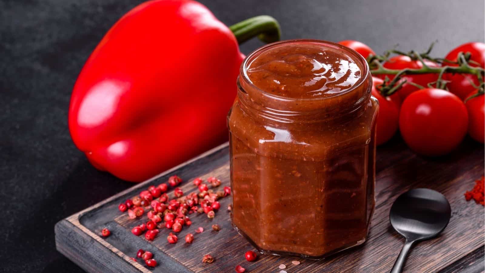 A jar of red sauce sits on a wooden board next to a red bell pepper, cherry tomatoes on the vine, scattered peppercorns, and a metal spoon.