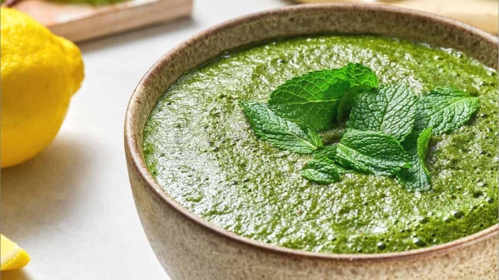 A bowl of green chutney garnished with fresh mint leaves, with a lemon next to it on a white surface.