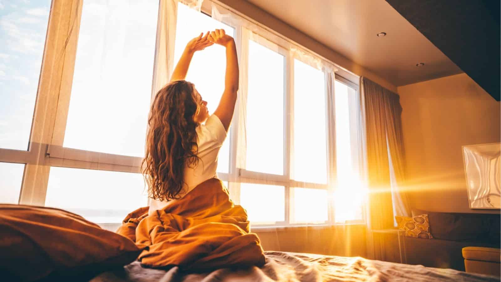 A person with long hair sits on a bed, stretching arms overhead, while sunlight streams through large windows in a modern bedroom.