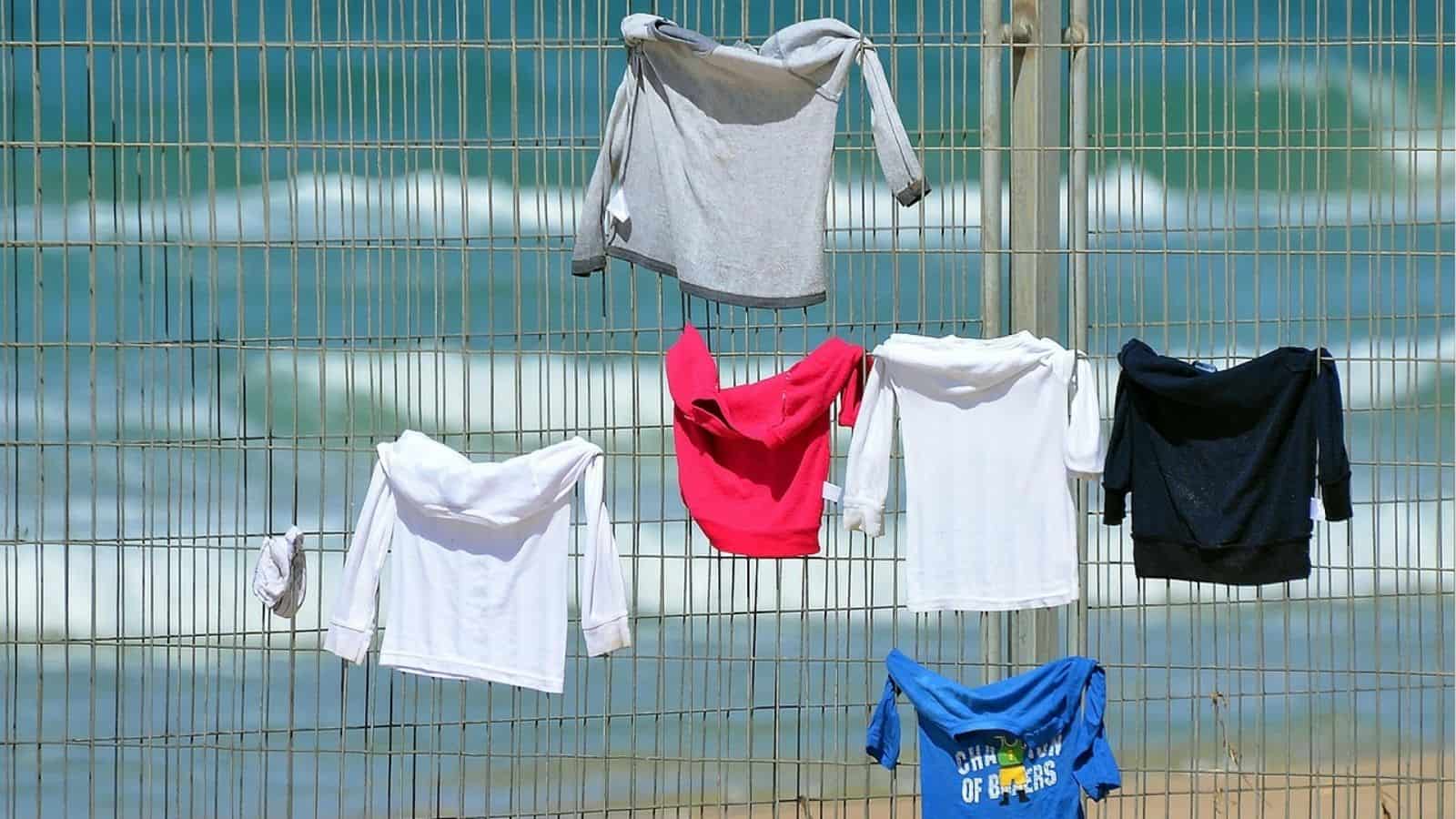 Six shirts in various colors are hung on a metal fence to dry, with an out-of-focus beach and ocean in the background.