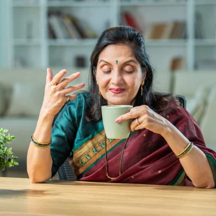 A woman in a saree sits at a table, holding a cup and gesturing with her other hand, with a plant and a blurred living room in the background.