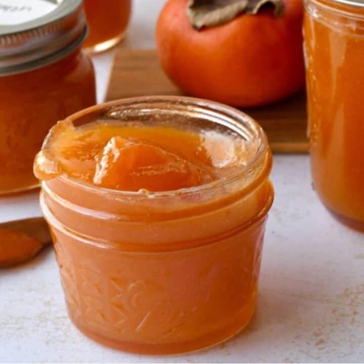 Small glass jars filled with orange persimmon jam, one open with jam and a spoon beside it, and a whole persimmon fruit in the background.